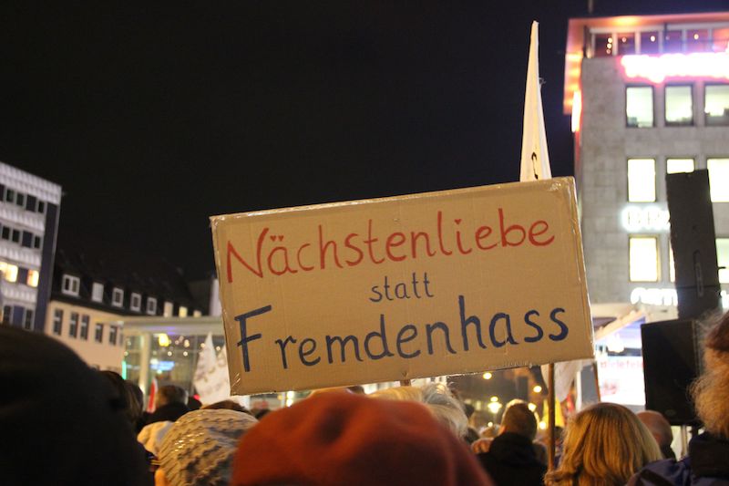 Bild einer abendlichen Demonstration gegen Pegida, man sieht ein Schild mit der Aufschrift "Nächstenliebe statt Fremdenhass", das in die Höhe gestreckt wird.