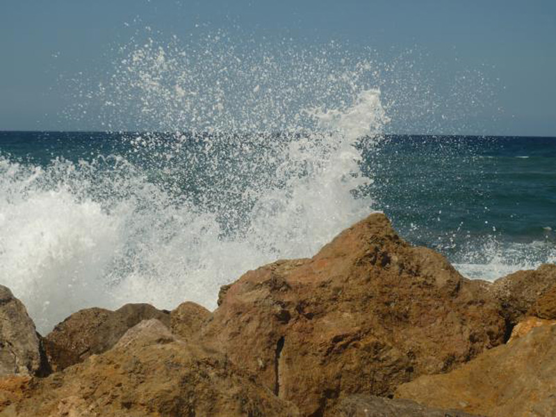 Eine Welle bricht sich am felsigen Ufer, das Wasser spritzt hoch.