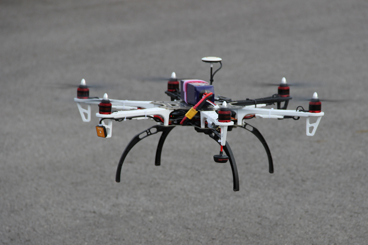 Bild einer kleinen im Flug befindlichen Drohne mit Videofunktion, im Hintergrund Asphalt.