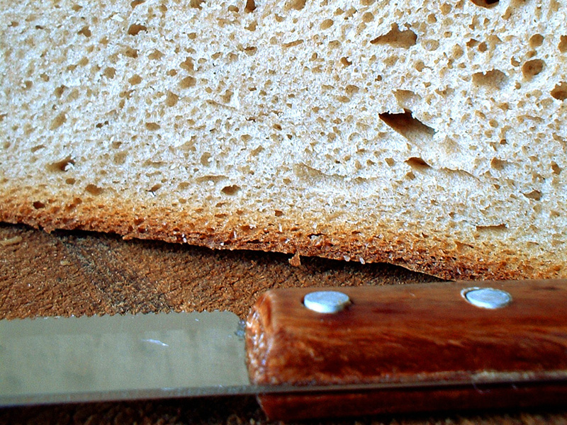 Nahaufnahme eines hellen Brotes auf einem Holzteller, vor dem Brot liegt ein Messer mit Holzgriff.