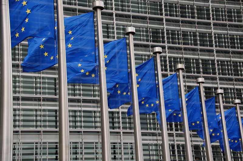 Bildausschnitt mehrerer EU-Flaggen, die an Fahnenmästen wehen, im Hintergund ein Gebäude der EU-Kommission in Brüssel.