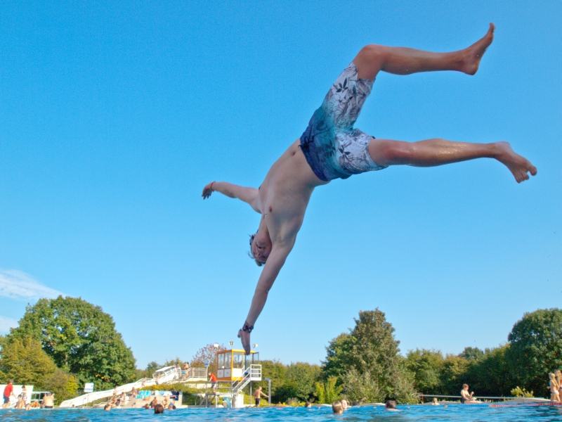 Bild eines Jungen, der bei strahlend blauem Himmel kopfüber in das Becken eines Freibades springt.