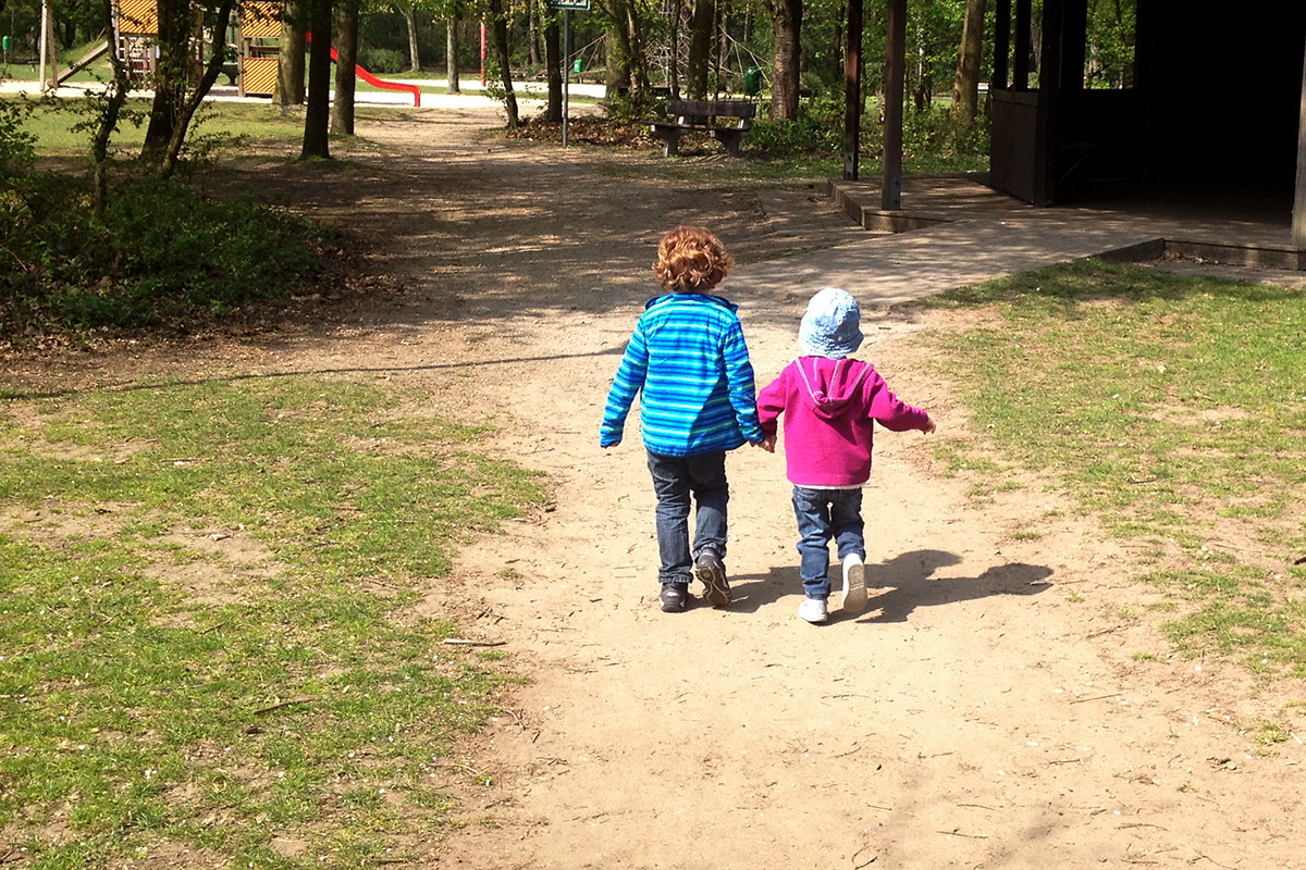 Zwei kleine Kinder laufen bei einem Spaziergang Hand in Hand vor dem Fotografierenden her in Richting Wald.