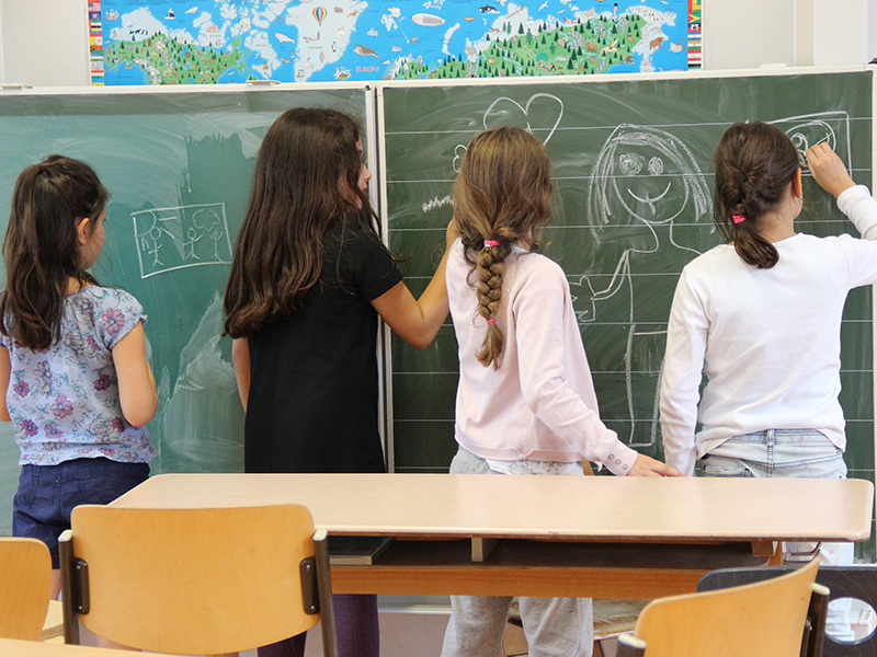 Vier Mädchen stehen an einer Tafel im Klassenzimmer und beschreiben und bemalen diese.
