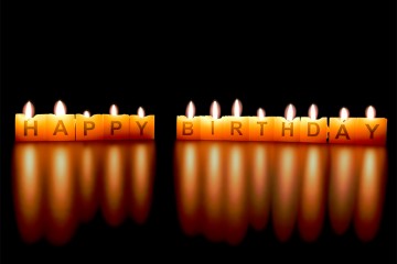 Vor dunklem Hintergrund stehen in einer Reihe kleine eckige Kerzen, die leuchten. Auf jeder Kerze steht ein Buchstabe, zusammen ergeben sie den Schriftzug "Happy Birthday".
