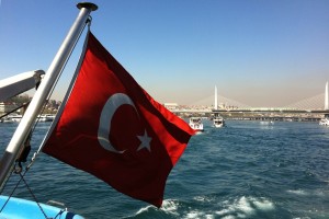 Türkische Staatsflagge am Bug eines Schiffes, mit Meer und Brücke im Hintergrund.