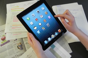 Weibliche Hände halten ein iPad und tippen darauf, im Hintergund liegen verschiedene Unterlagen.