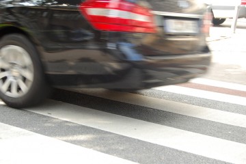 Bildausschnitt: man sieht das Heck eines schwarzen Autos, das offensichtlich zu schnell über einen Zebrastreifen fährt.