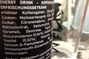Nahaufnahme einer Dose Energy-Drink, man sieht einen Teil der Zutatenliste in weißer Schrift auf schwarzen Grund, im Hintergund stehen Gläser.