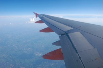 Blick aus dem Flugzeugfenster auf die Tragfläche, man sieht im Hintergrund etwas Landschaft, eine Wolke und den Himmel.