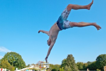 Bild eines Jungen, der bei strahlend blauem Himmel kopfüber in das Becken eines Freibades springt.