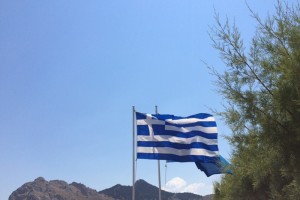 Eine griechische Flagge weht im Wind, im Hintergrund ist hügeliges Gelände und blauer Himmel zu sehen.