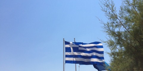 Eine griechische Flagge weht im Wind, im Hintergrund ist hügeliges Gelände und blauer Himmel zu sehen.