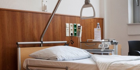 Krankenhausbett mit aufgeschlagener weißer Bettdecke vor einer Holzpaneele an der Wand, auf dem Beistelltisch steht eine Flasche Wasser.