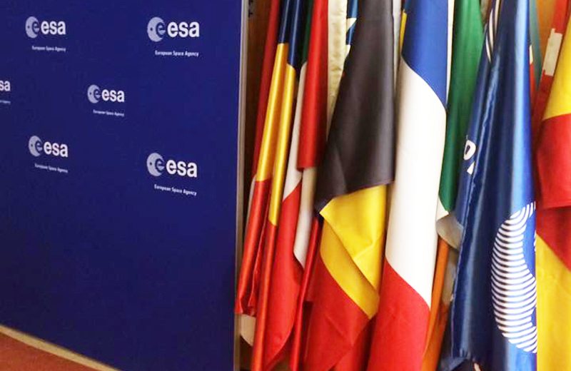 Verschiedene europäische Landerflaggen sowie eine Falle der eruopäischen Weltraumbehörde ESA, die neben einer dunkelblauen, mit dem ESA-Logo versehenen Wand aufgestellt wurden